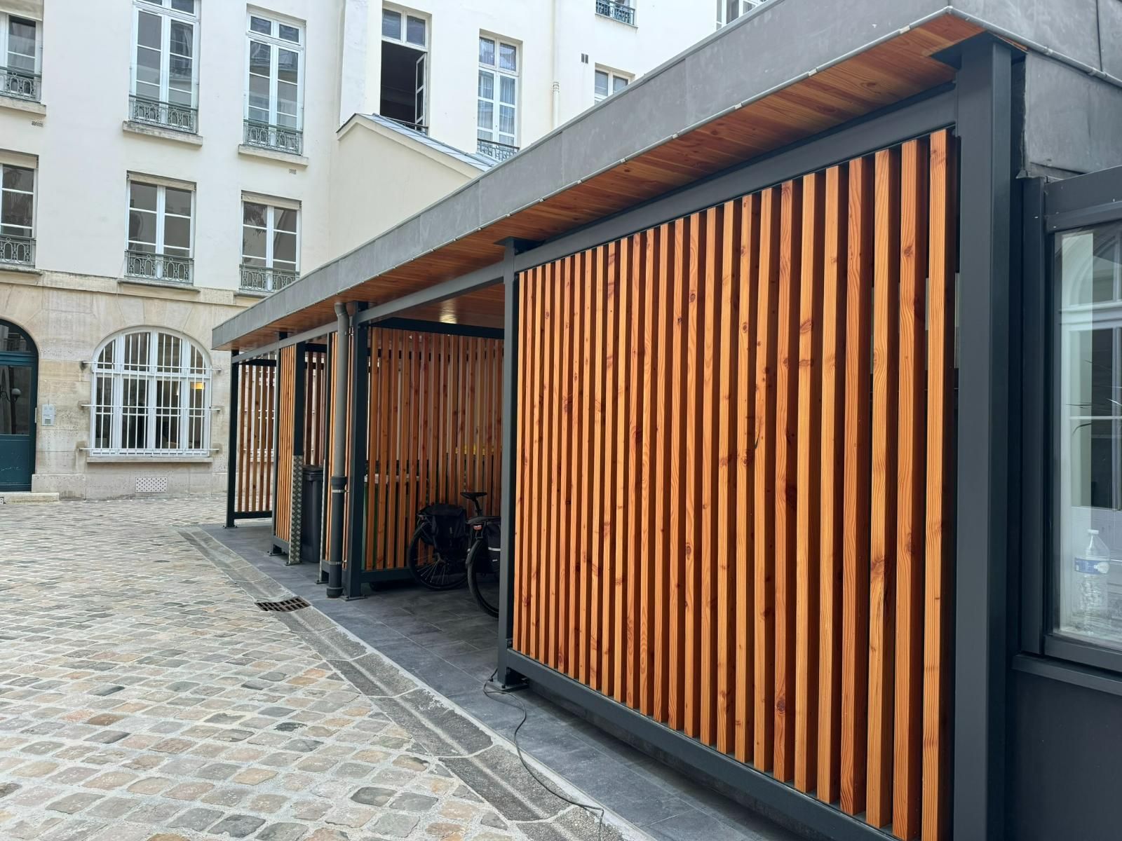 Abri à vélos en bois à lattes verticales, sous un toit gris, dans une cour pavée.