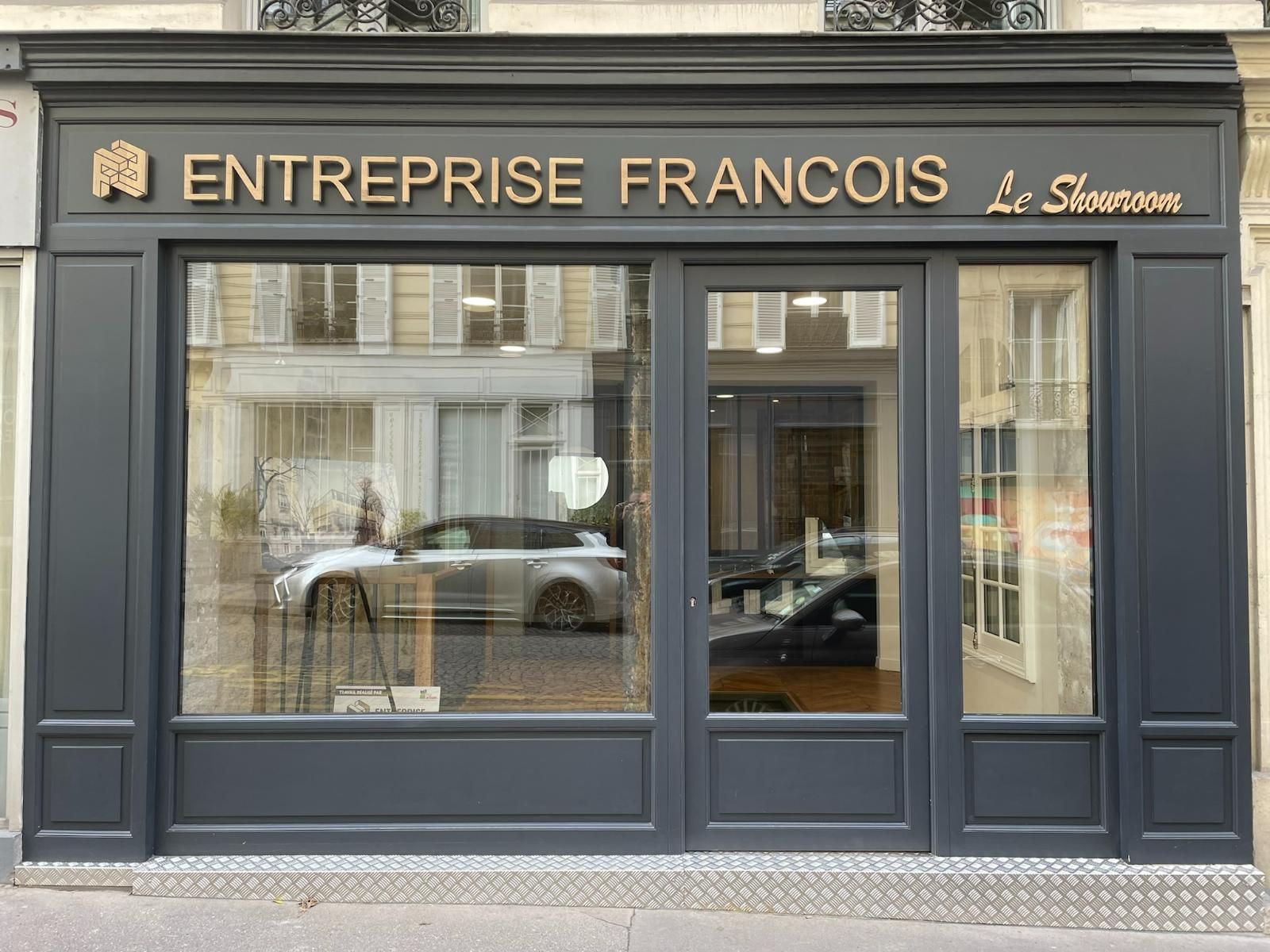 Façade de magasin gris foncé avec inscription dorée : « ENTREPRISE FRANÇAIS Le Showroom ». Vitrine réfléchissante présentant une voiture.