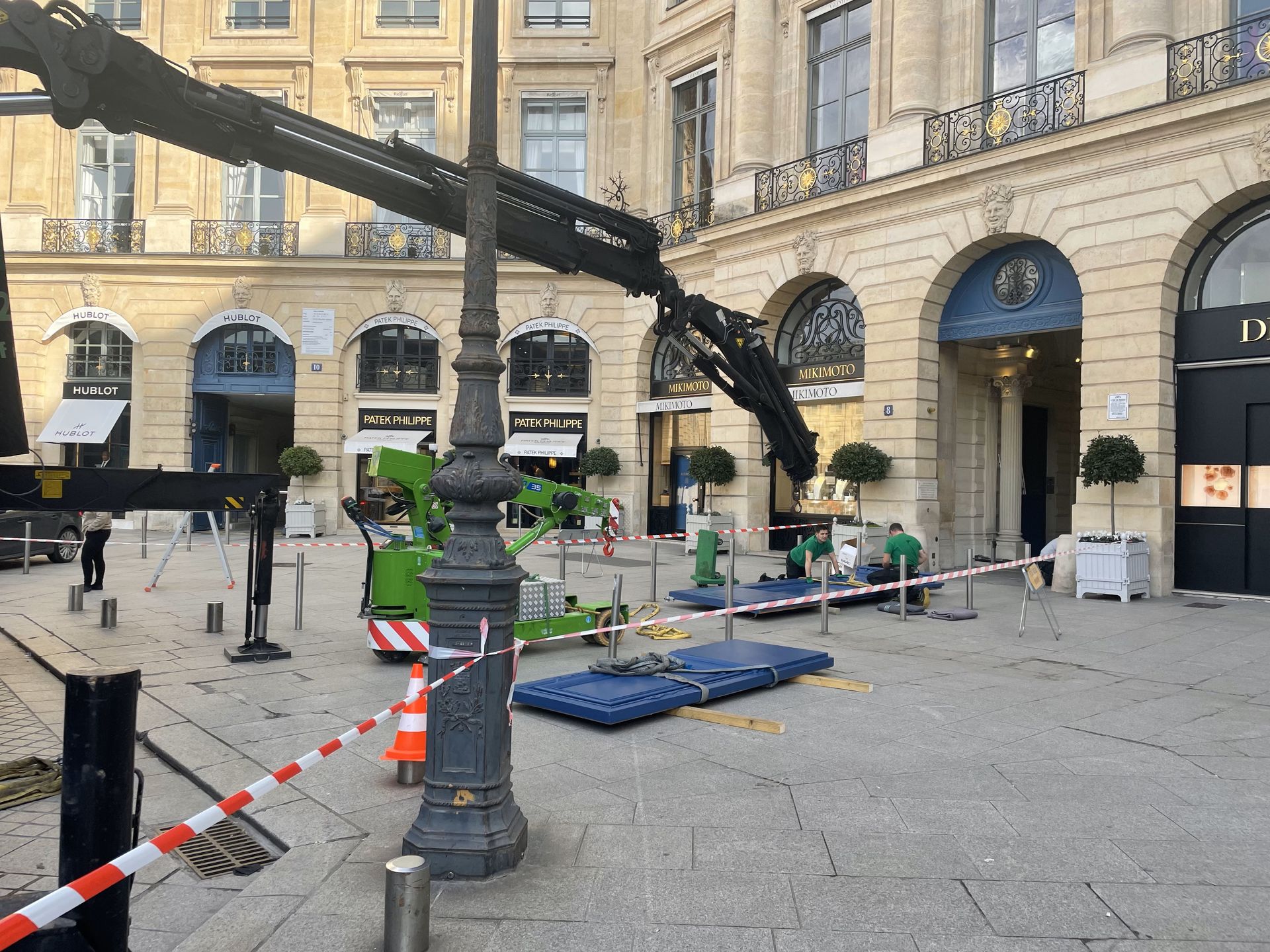 Une grue de chantier et son matériel sont installés sur une place pavée devant un immeuble orné d'arches à Paris.