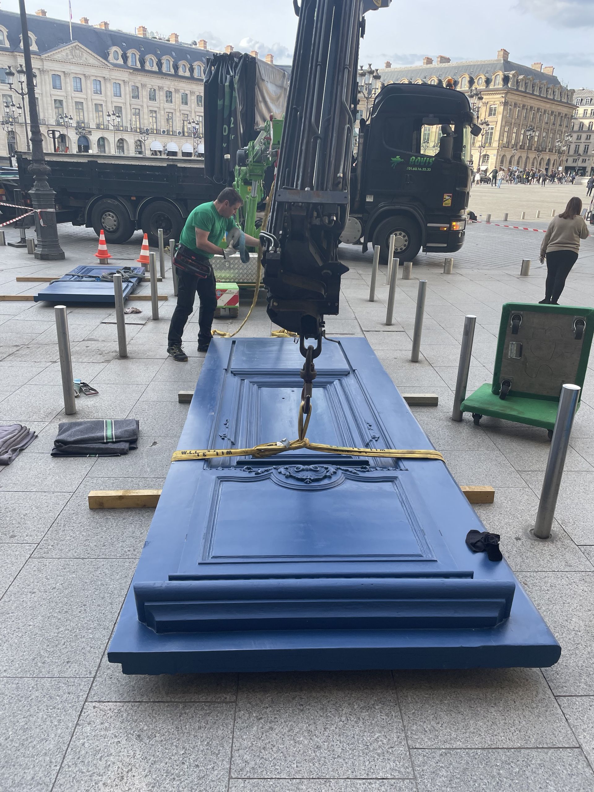 Sur une place historique de la ville, une grue soulève un grand panneau de porte rectangulaire bleu roi tandis que des ouvriers se préparent à l'installer.