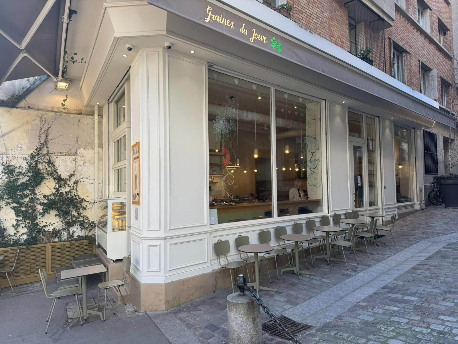 Café d'angle avec façade blanche, grandes fenêtres, terrasse sur rue pavée et mur recouvert de lierre.