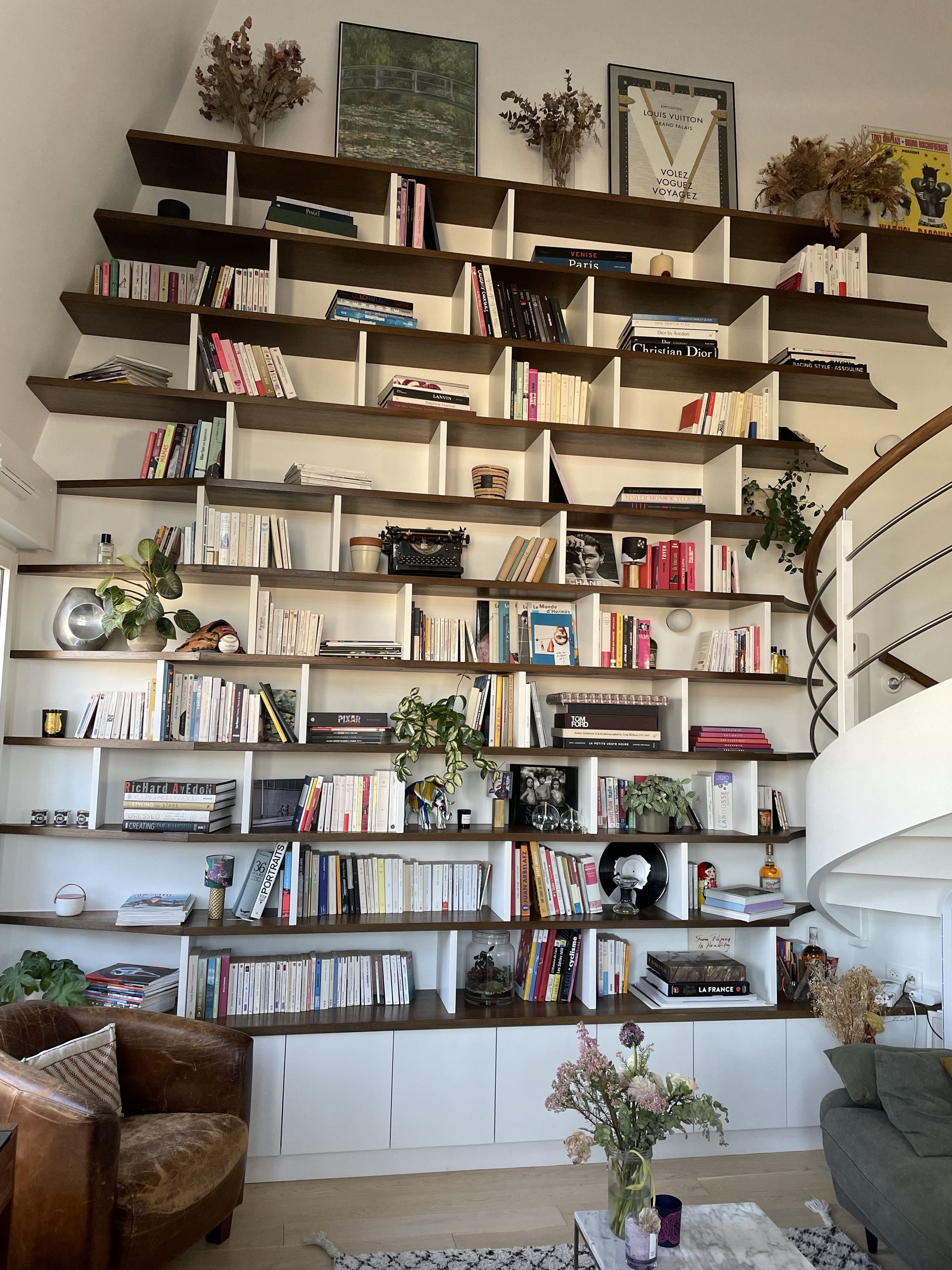 Une imposante bibliothèque murale à plusieurs niveaux, remplie de livres, de plantes et d'objets décoratifs, se trouve à côté d'un escalier en colimaçon.