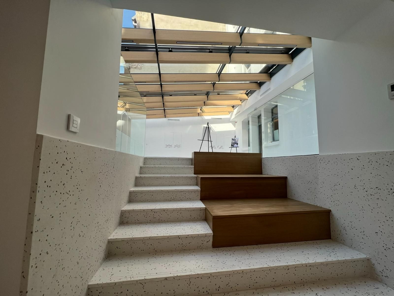 Escalier aux marches en terrazzo blanc et à plateforme en bois menant à un espace ouvert et ensoleillé avec un plafond partiellement vitré.