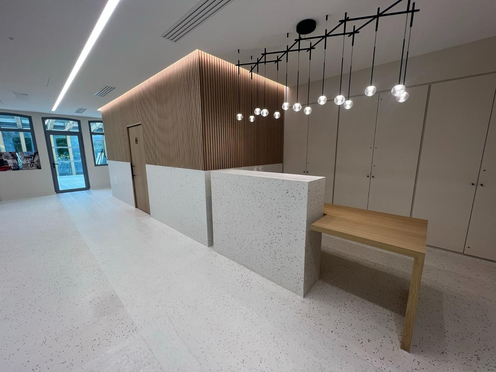 Un comptoir d'accueil moderne doté d'un socle style terrazzo, de panneaux de bois et d'un luminaire suspendu linéaire artistique.
