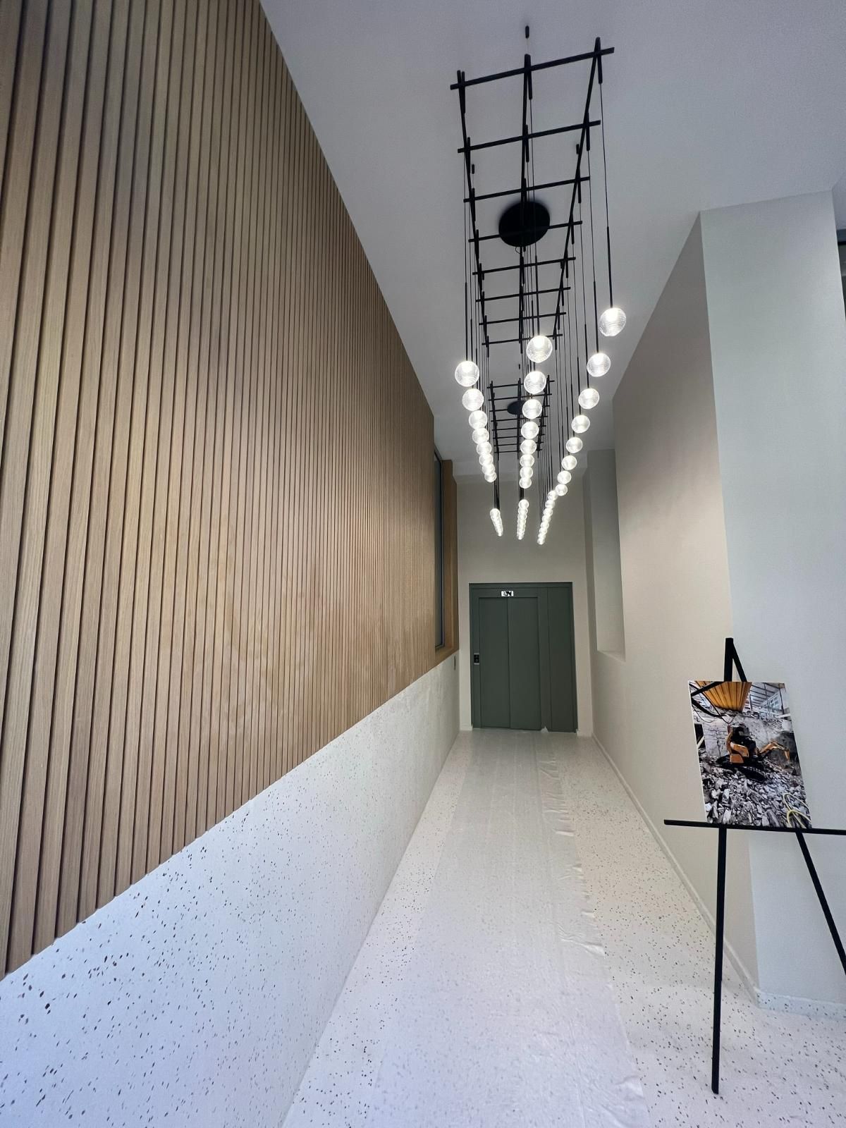 Long couloir avec mur lambrissé de bois clair, sol blanc moucheté, luminaire moderne et œuvre d'art sur chevalet.