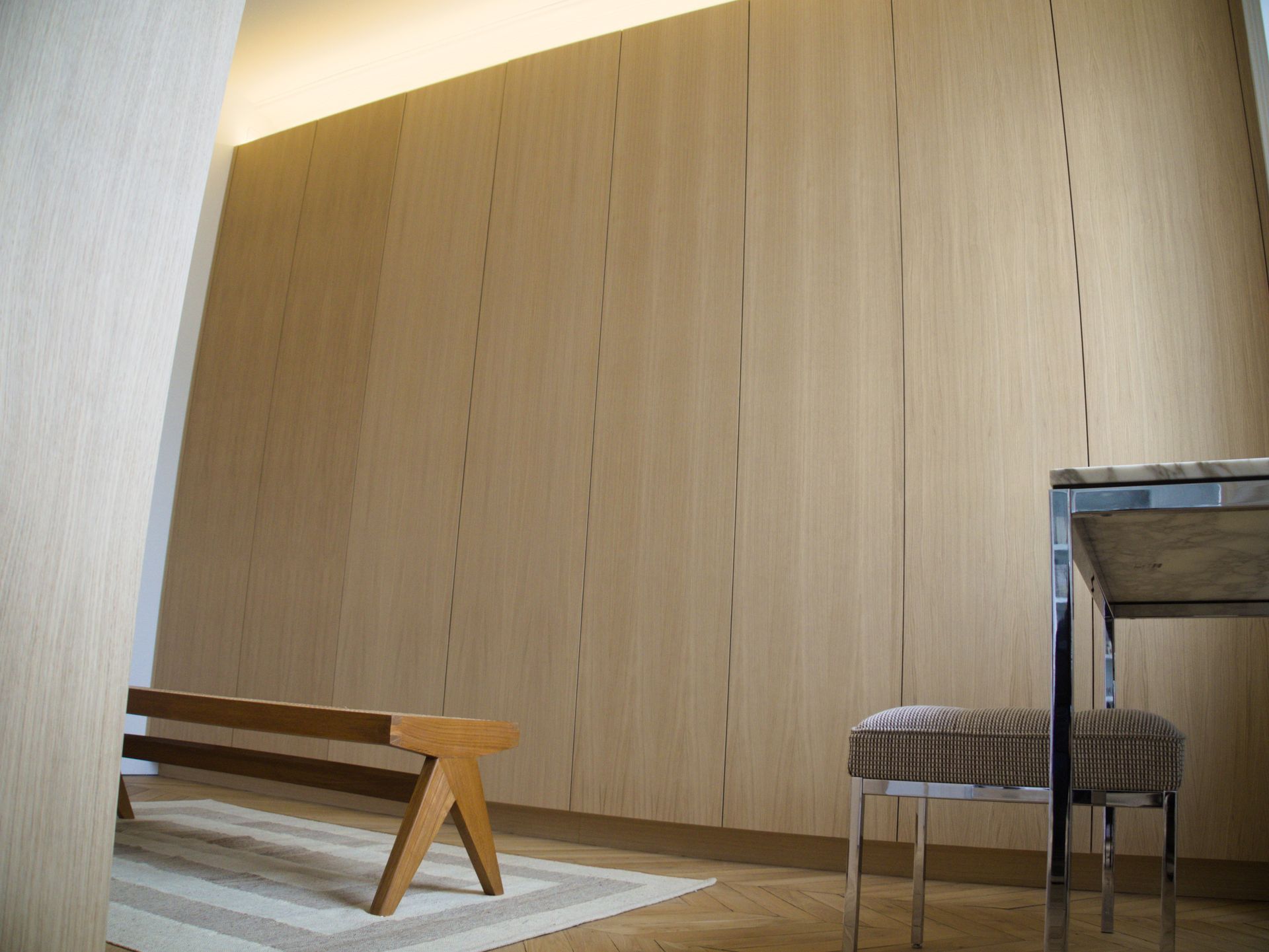 Un mur lambrissé de bois clair habille une pièce où se trouvent un banc bas en bois posé sur un tapis et une petite table avec un tabouret à motifs à proximité.