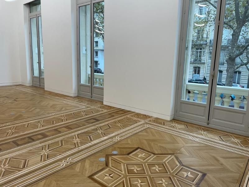 Une pièce lumineuse et vide, dotée d'un parquet en bois ouvragé et de grandes portes-fenêtres à cadre blanc donnant sur une rue.