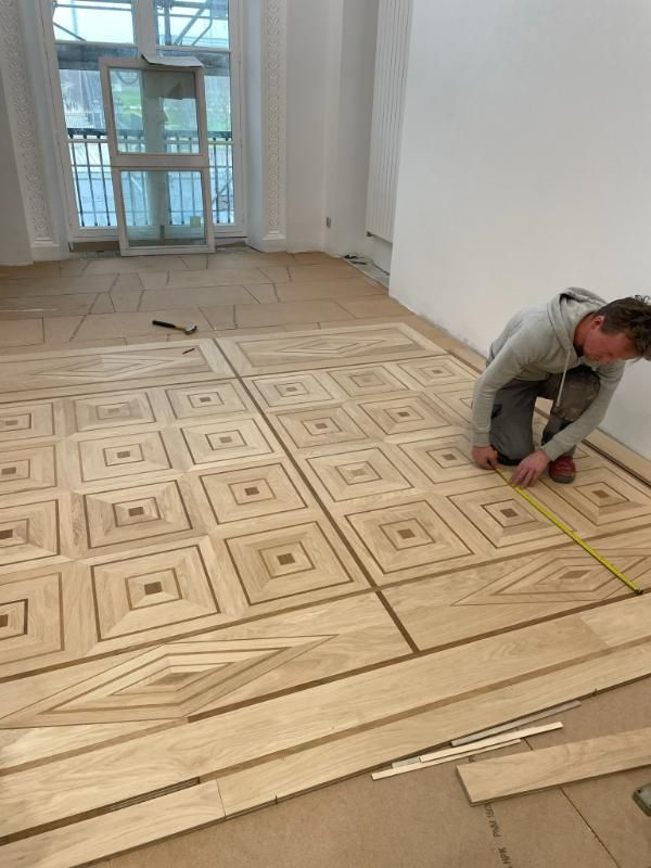 Une personne s'agenouille pour mesurer un parquet en bois clair aux motifs géométriques complexes, en cours d'installation dans une pièce dotée d'une grande fenêtre.