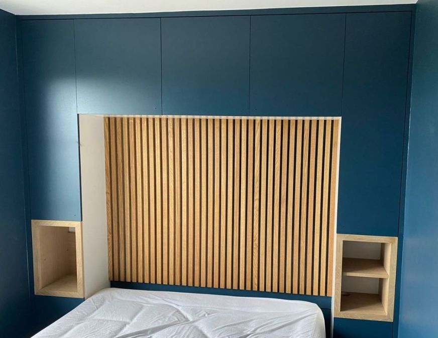 Un mur de chambre orné de placards intégrés bleu marine encadrant une tête de lit centrale à lattes verticales en bois avec des alcôves latérales.
