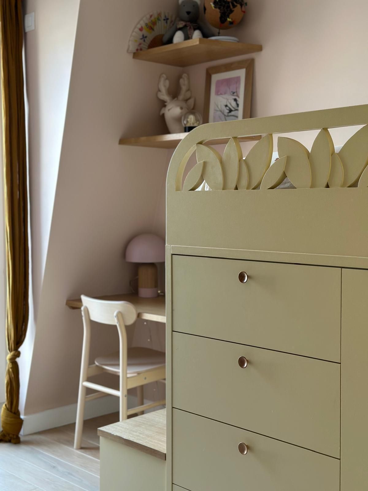 Chambre d'enfant avec murs roses, meubles en bois clair, bureau, étagères et une armoire jaune-vert ornée de motifs de feuilles.