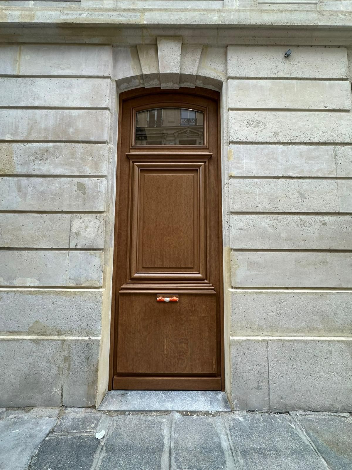 Une porte en bois brun avec une petite fenêtre en partie supérieure, encastrée dans un mur de pierre orné de lignes horizontales en retrait.