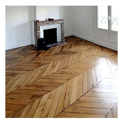 Une pièce vide avec un parquet clair à chevrons, une cheminée en marbre et des murs blancs.