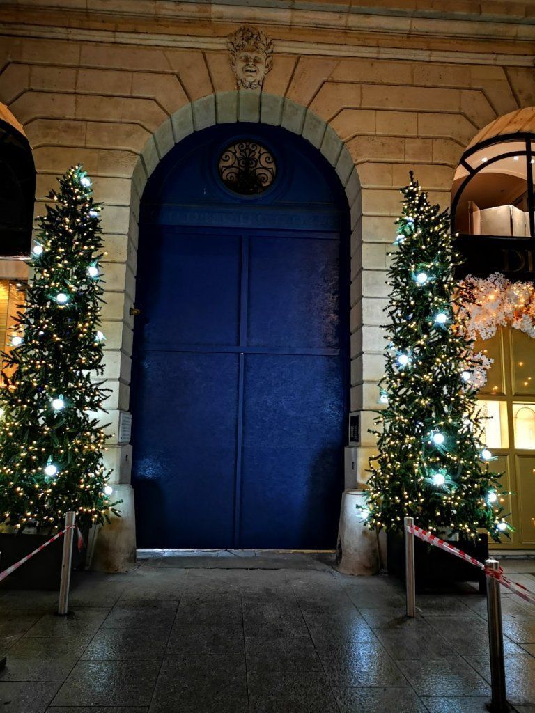 Une grande porte cintrée bleue, encastrée dans un mur de pierre, est flanquée de deux sapins de Noël décorés de guirlandes lumineuses.