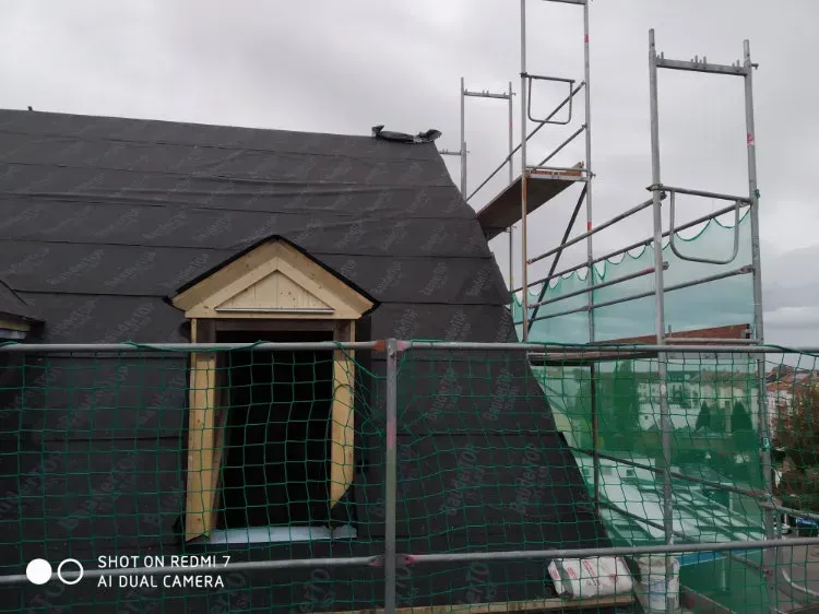 Zimmerei Andreas Karl GmbH - Dach eines Gebäudes mit Dachfenster, umgeben von Baugerüst mit grünem Fangnetz