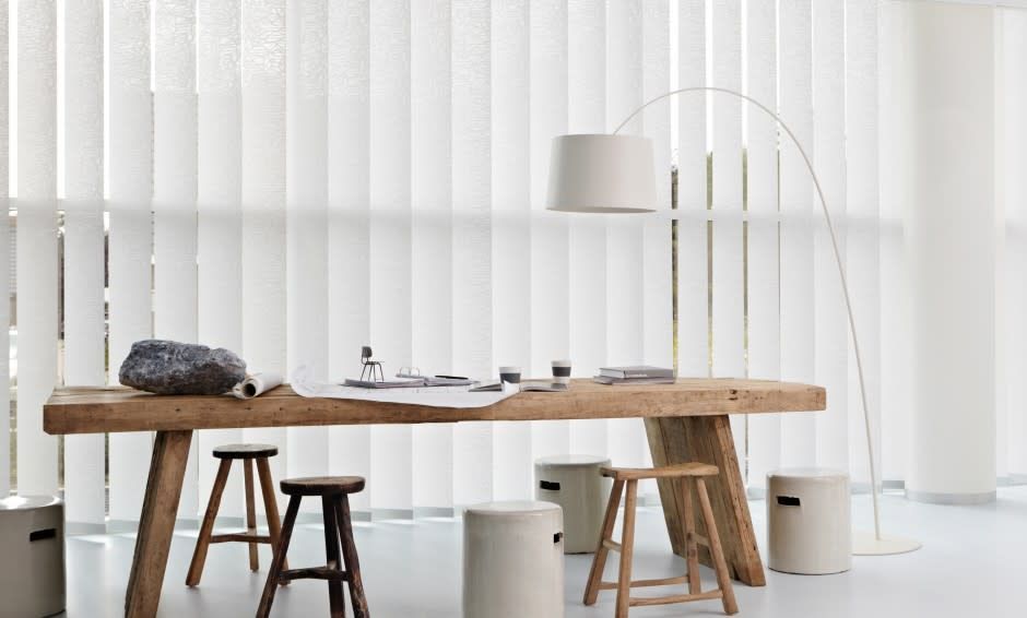 Un tavolo di legno con sgabelli e una lampada in una stanza con tende verticali bianche.