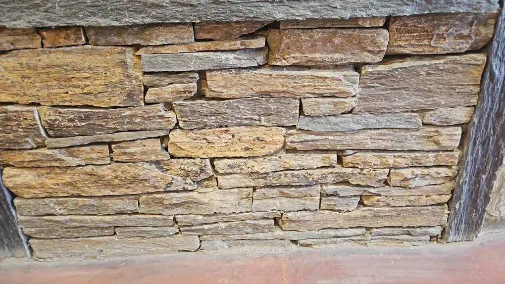 Muro de piedra con diferentes tonos de piedras marrones y grises, debajo una losa de piedra gris oscuro y vigas de madera.