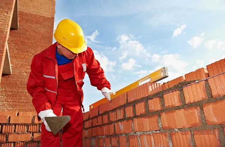 Albañil con mono rojo y casco amarillo usando un nivel para construir una pared de ladrillos contra un cielo azul.