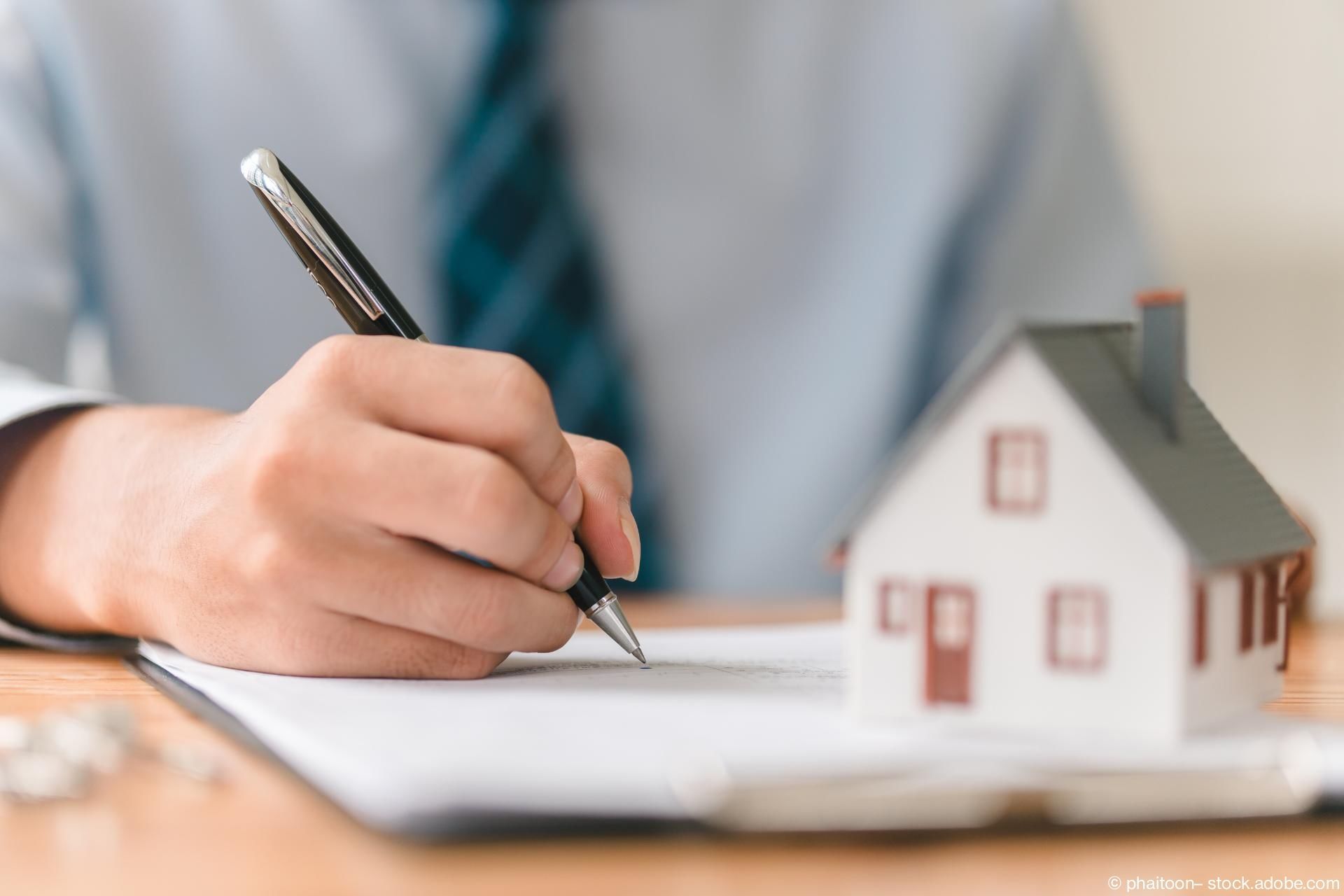 Hand mit Kugelschreiber schreibt auf Papier, daneben Modell eines Hauses mit Schornstein und Fenstern.