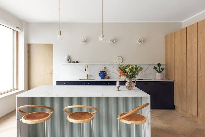 Una cocina moderna con una isla de mármol blanco, base de listones verde claro, tres taburetes de madera y armarios