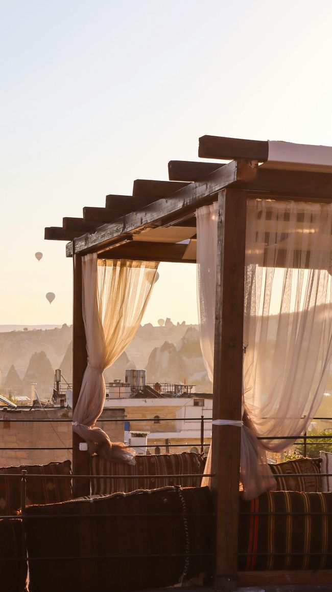 Una pérgola de madera con cortinas blancas y vaporosas ofrece vistas a un paisaje montañoso salpicado de globos aerostáticos