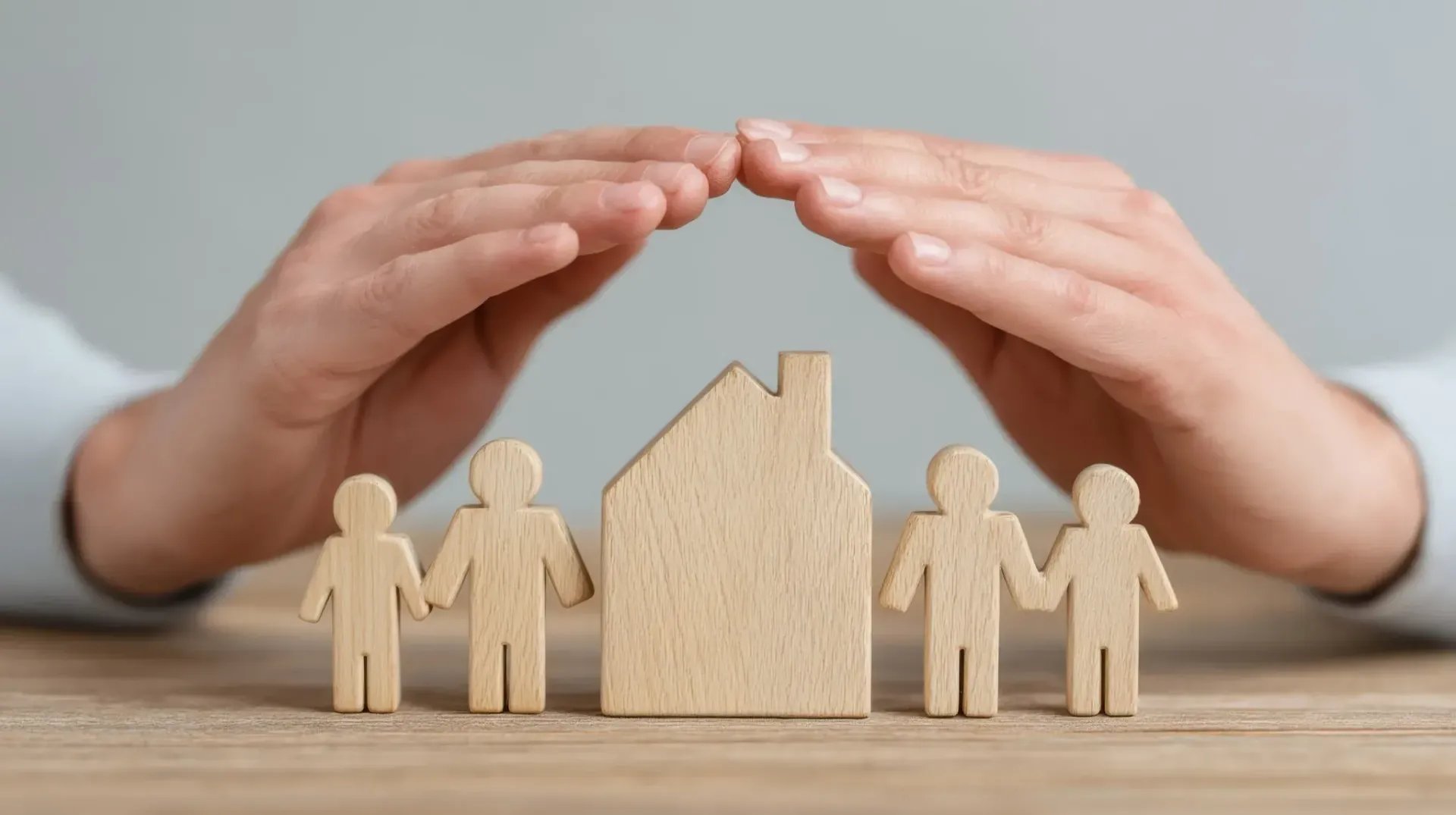 Manos protegiendo una casa de madera y figuras familiares, simbolizando la seguridad del hogar y la familia.