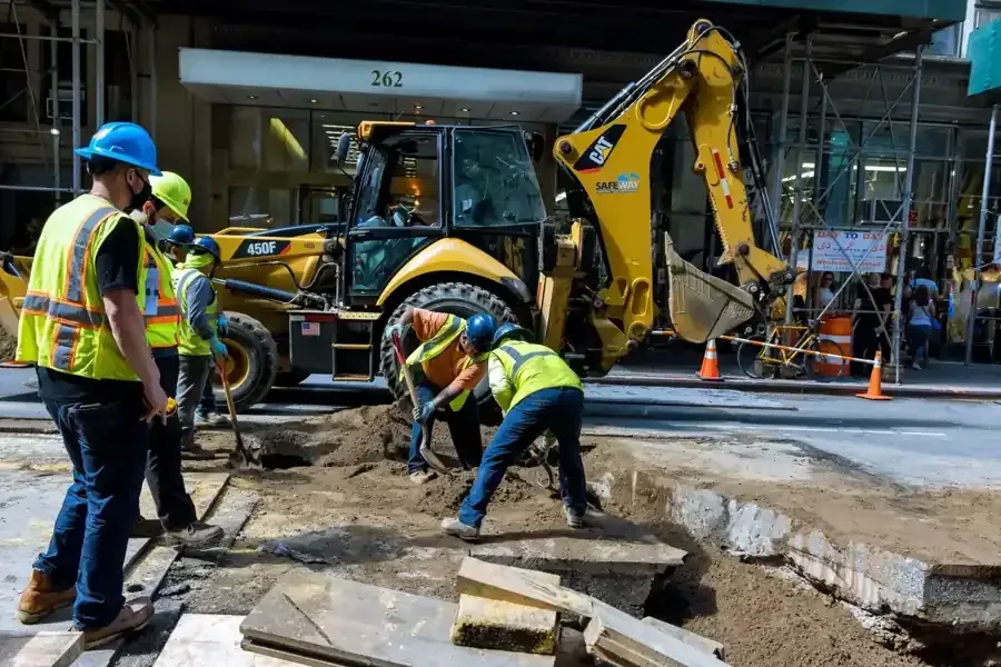 Obreros de la construcción utilizan una excavadora para cavar en una calle; llevan cascos y chalecos de seguridad.