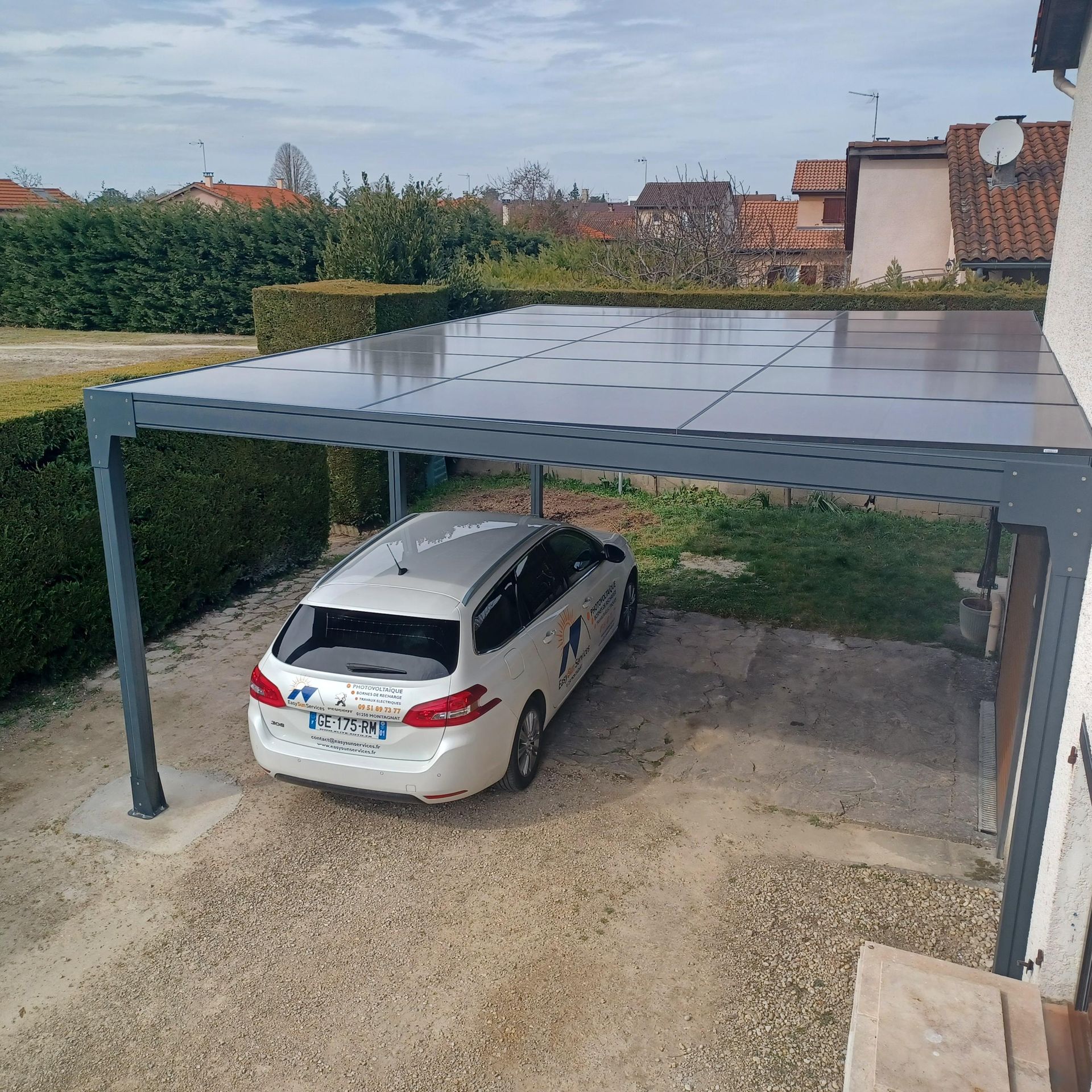 Voiture garée sous un abri équipé de panneaux solaires ; maisons et verdure en arrière-plan.