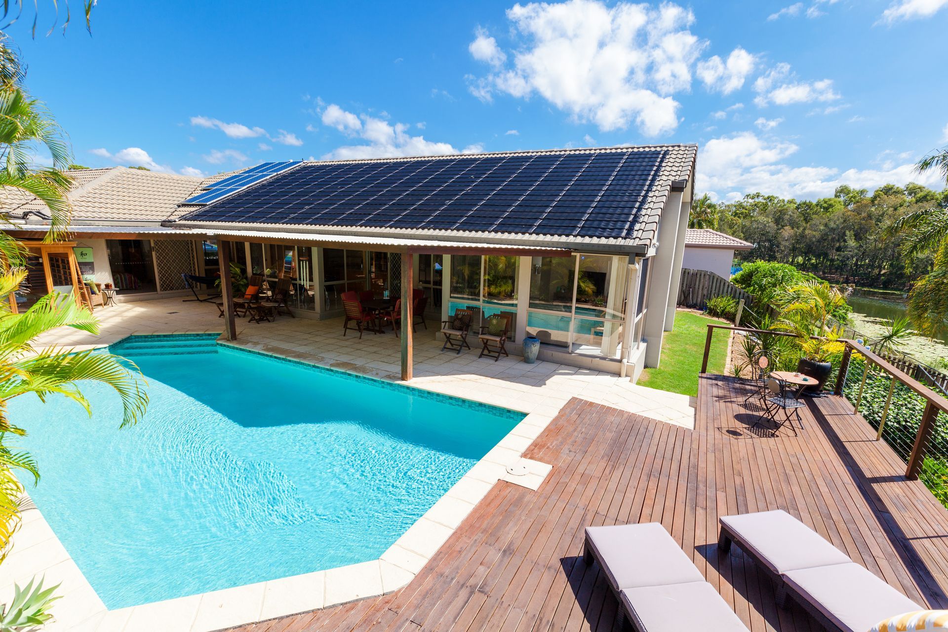 Maison avec piscine et terrasse en bois ; panneaux solaires sur le toit, arbres en arrière-plan.