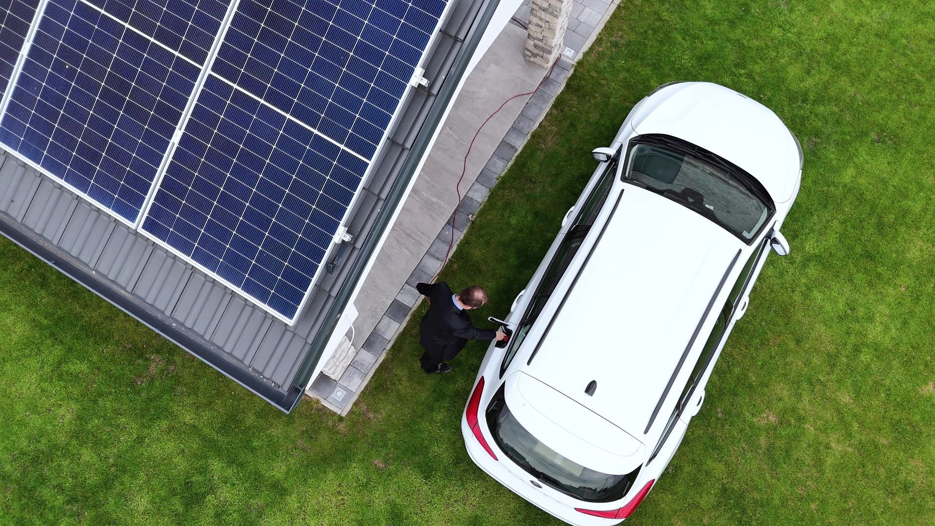 Panneaux solaires sur le toit d'une maison à côté d'une voiture blanche, une personne près de la voiture sur l'herbe verte.
