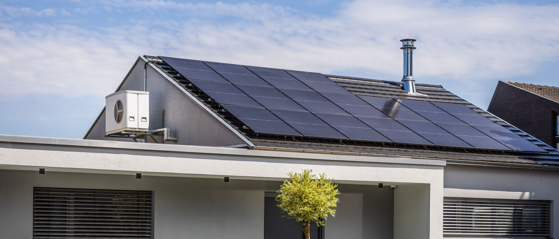 Une maison avec des panneaux solaires sur le toit, une cheminée et un petit arbre devant.