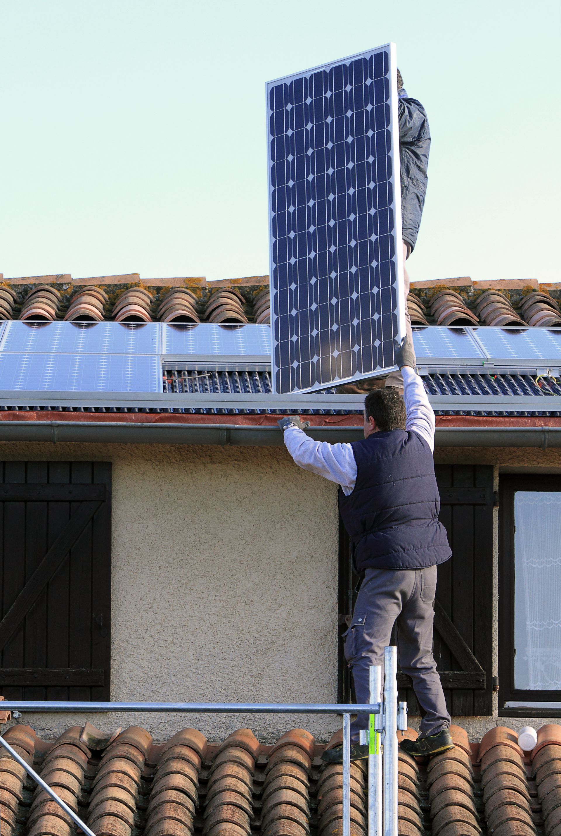 Des ouvriers installent un panneau solaire sur un toit de tuiles, à l'aide d'un échafaudage.