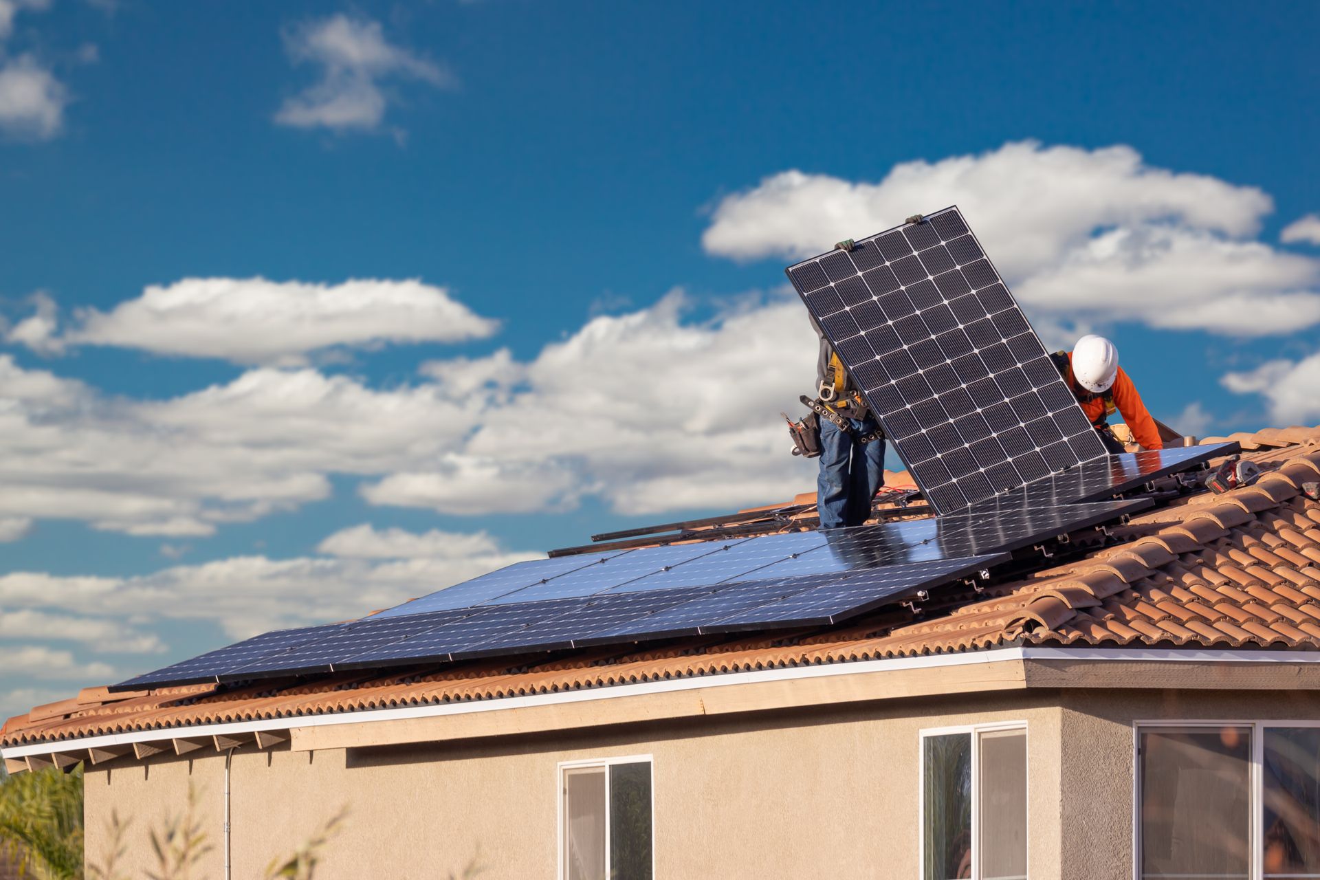Des ouvriers installent des panneaux solaires sur le toit d'une maison, sous un ciel bleu nuageux.