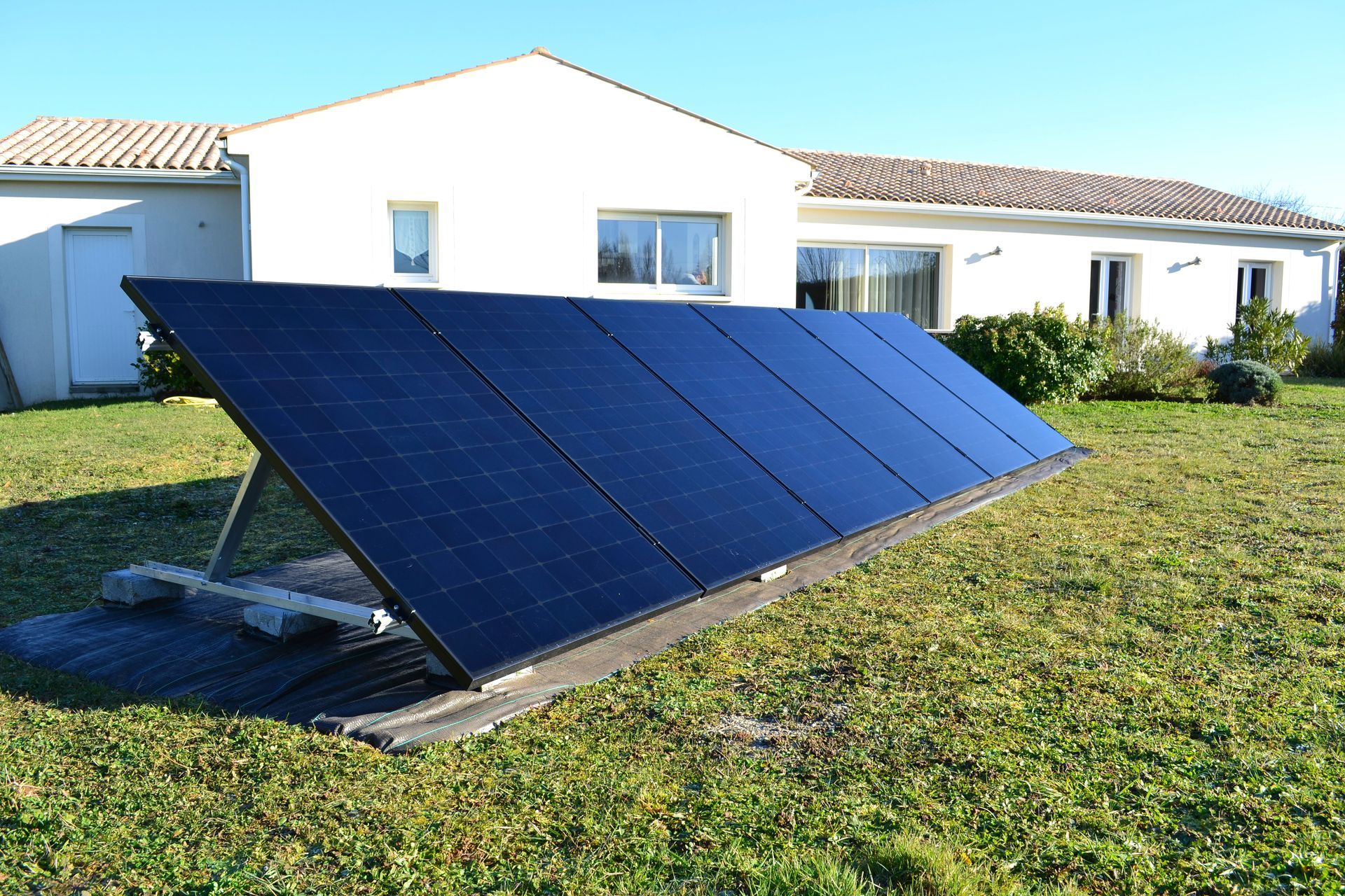 Des panneaux solaires sur un support devant une maison blanche par une journée ensoleillée.