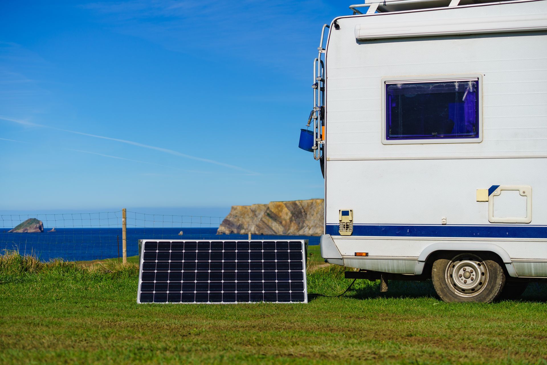 Panneau solaire sur l'herbe à côté d'une caravane avec vue sur l'océan sous un ciel bleu.
