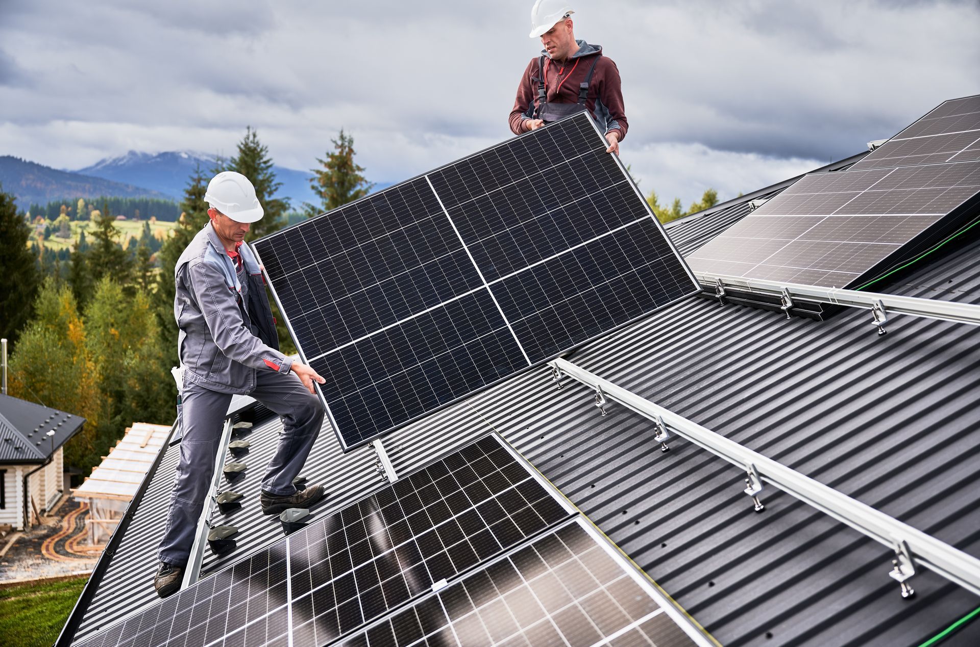 Deux ouvriers portant des casques de chantier installent des panneaux solaires sur un toit avec une montagne en arrière-plan.
