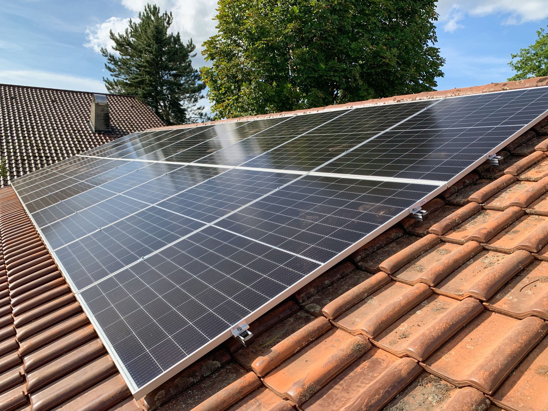 Des panneaux solaires installés sur un toit de tuiles rouges se détachent sur un ciel bleu, avec des arbres en arrière-plan.