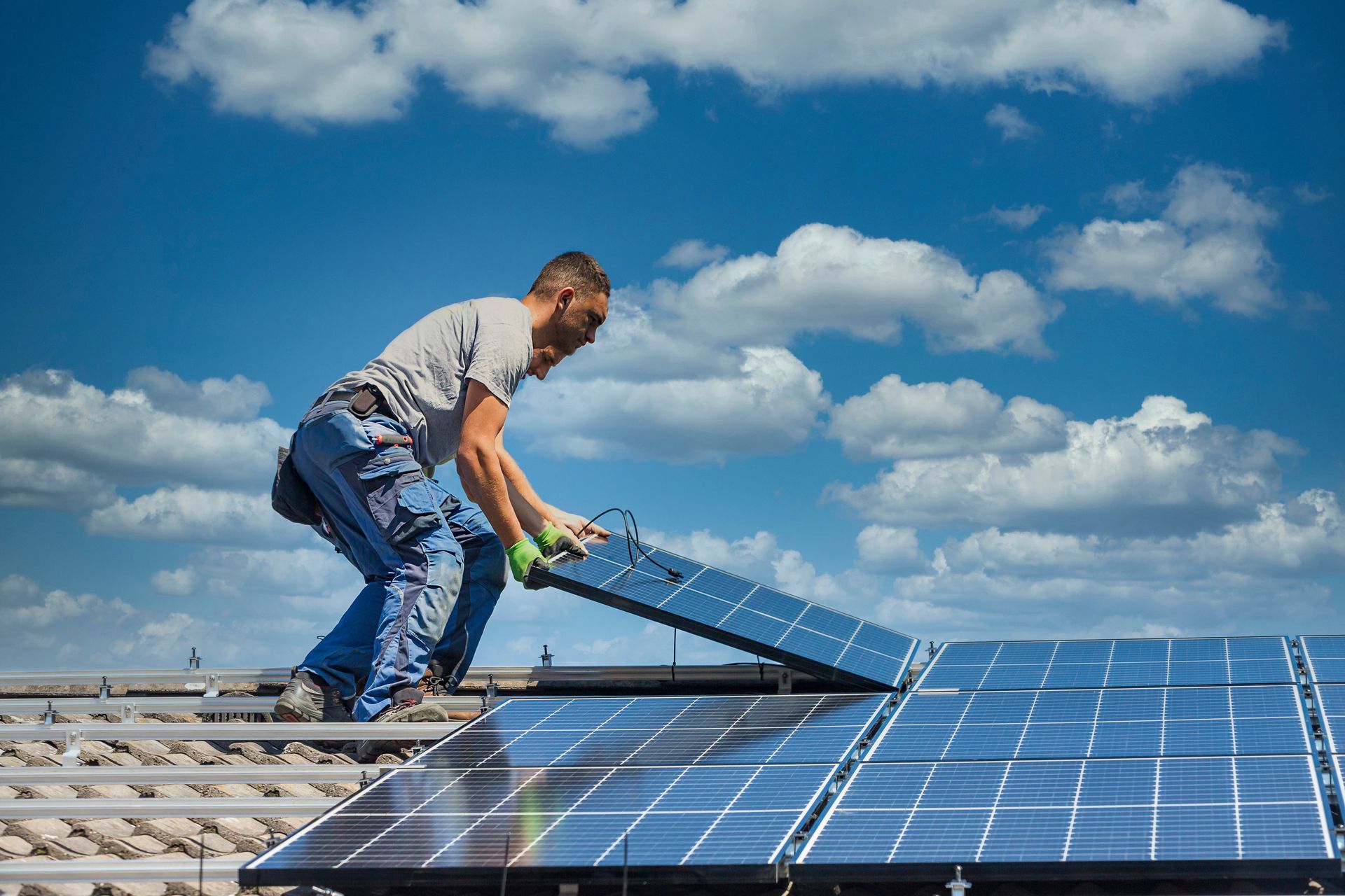 Un homme installe un panneau solaire sur un toit sous un ciel bleu nuageux.