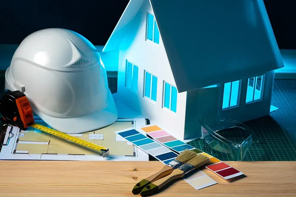 Un casco, una cinta métrica, pinceles y una maqueta de casa sobre una mesa.