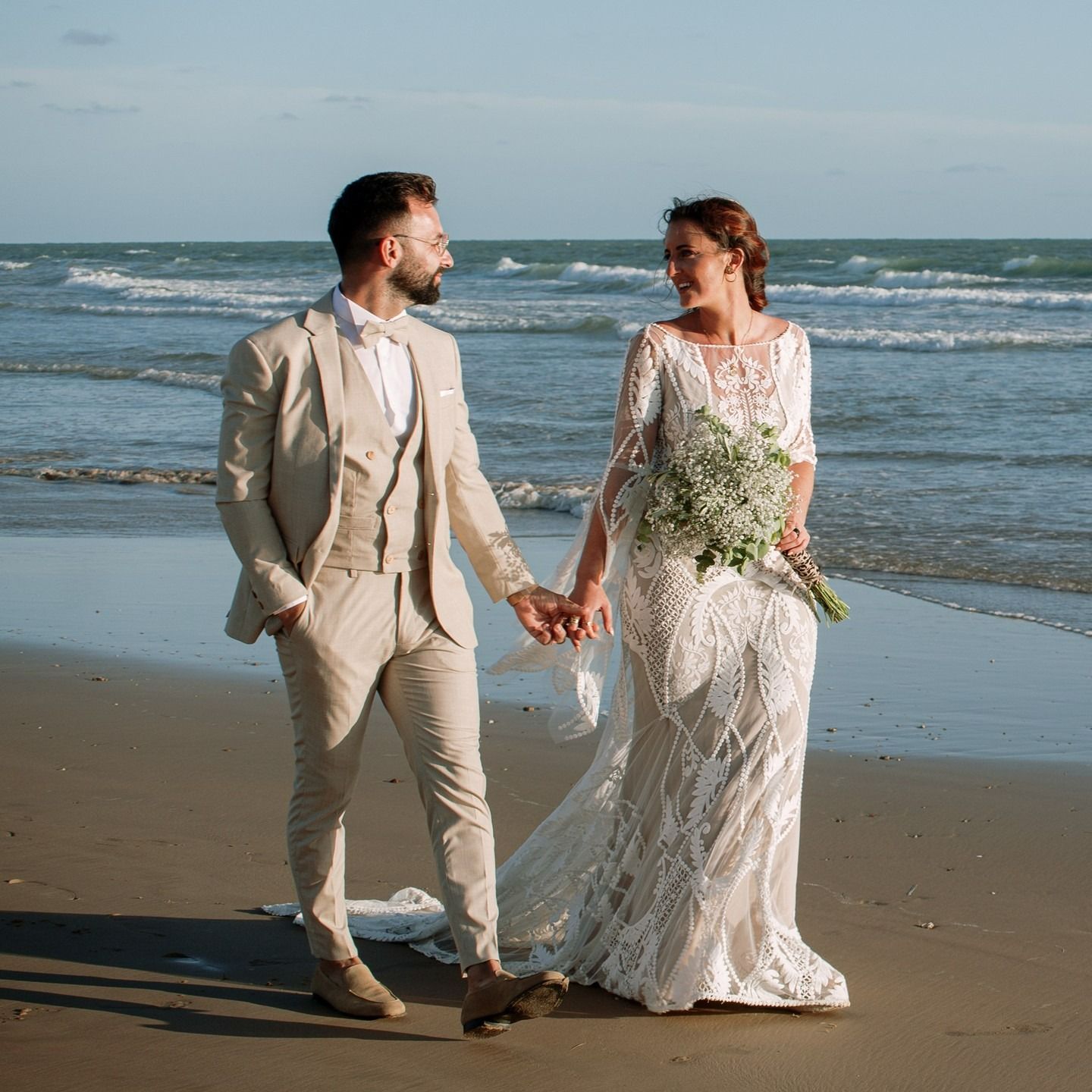 Una novia y un novio caminan por la playa tomados de la mano.
