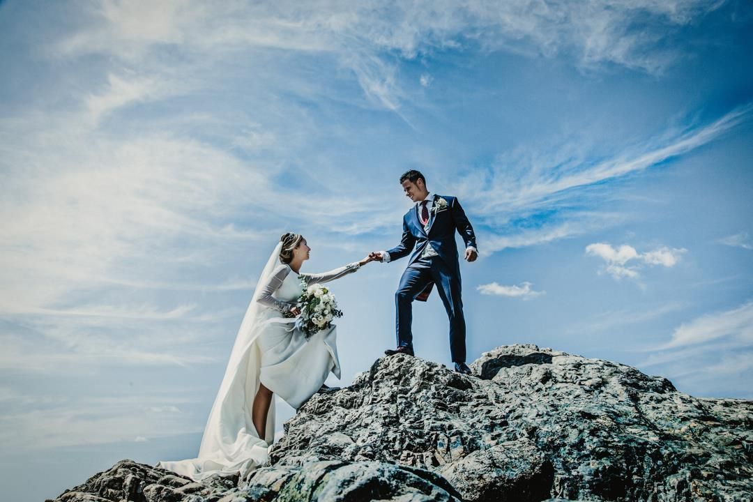 Una novia y un novio están de pie en la cima de una colina rocosa tomados de la mano.