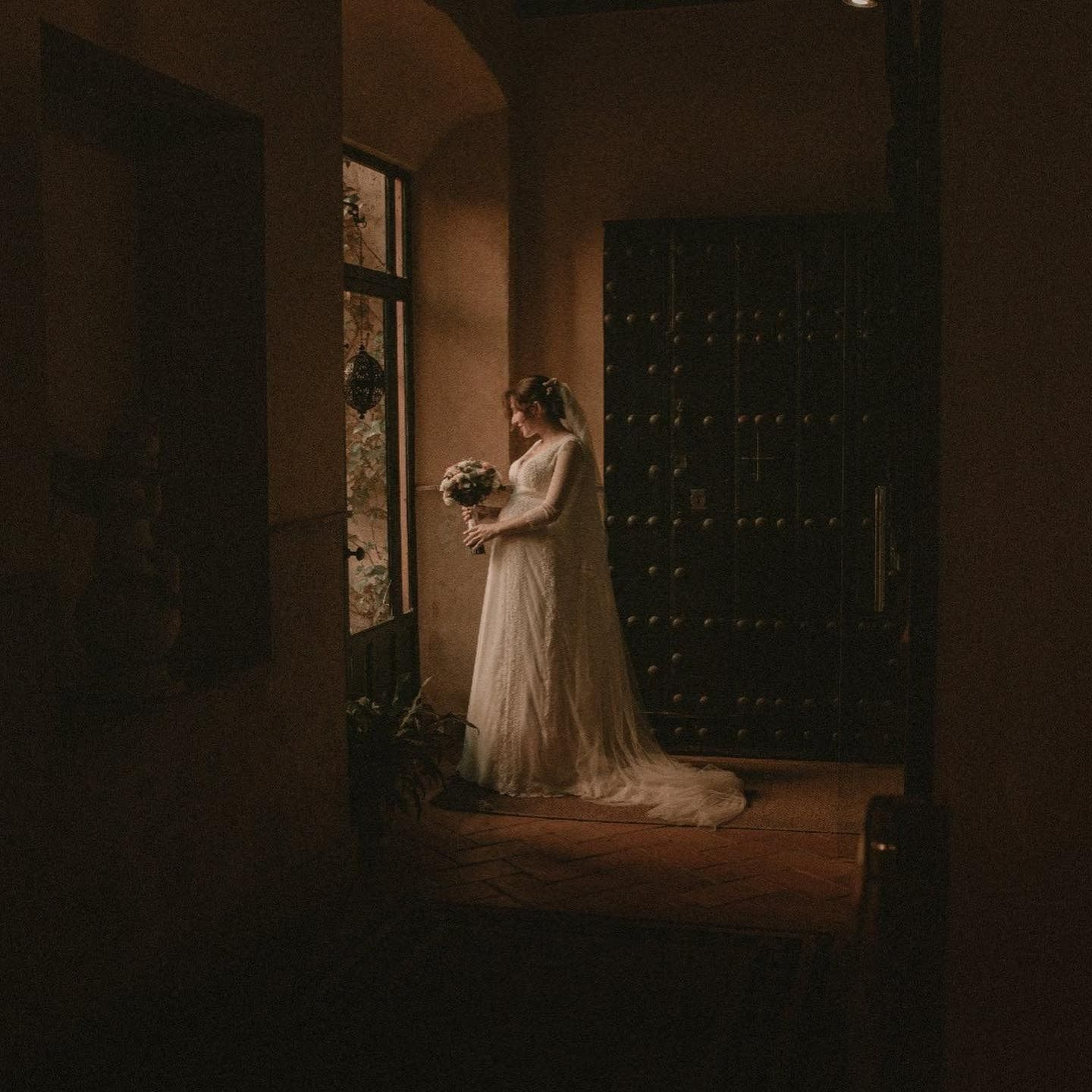 Una novia con un vestido de novia está parada frente a una ventana sosteniendo un ramo de flores.