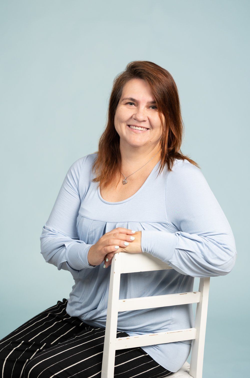 Hymyilevä nainen, jolla on yllään vaaleansininen pitkähihainen paita ja mustavalkoraidalliset housut, istuu valkoisella tuolilla.