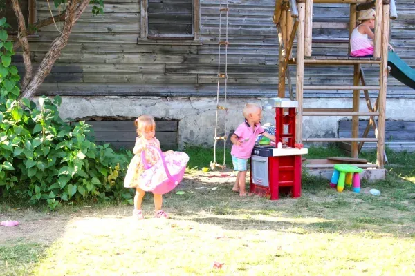 Kolme lasta leikkii takapihalla puurakennelman, leikkikeittiön ja pienen monivärisen jakkaran lähellä.