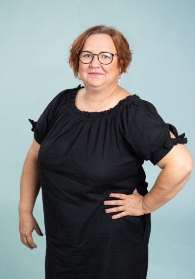 Mustaan ​​puhvihihaiseen ja silmälaseihin pukeutunut henkilö poseeraa käsi lantiolla vaaleansinistä taustaa vasten.