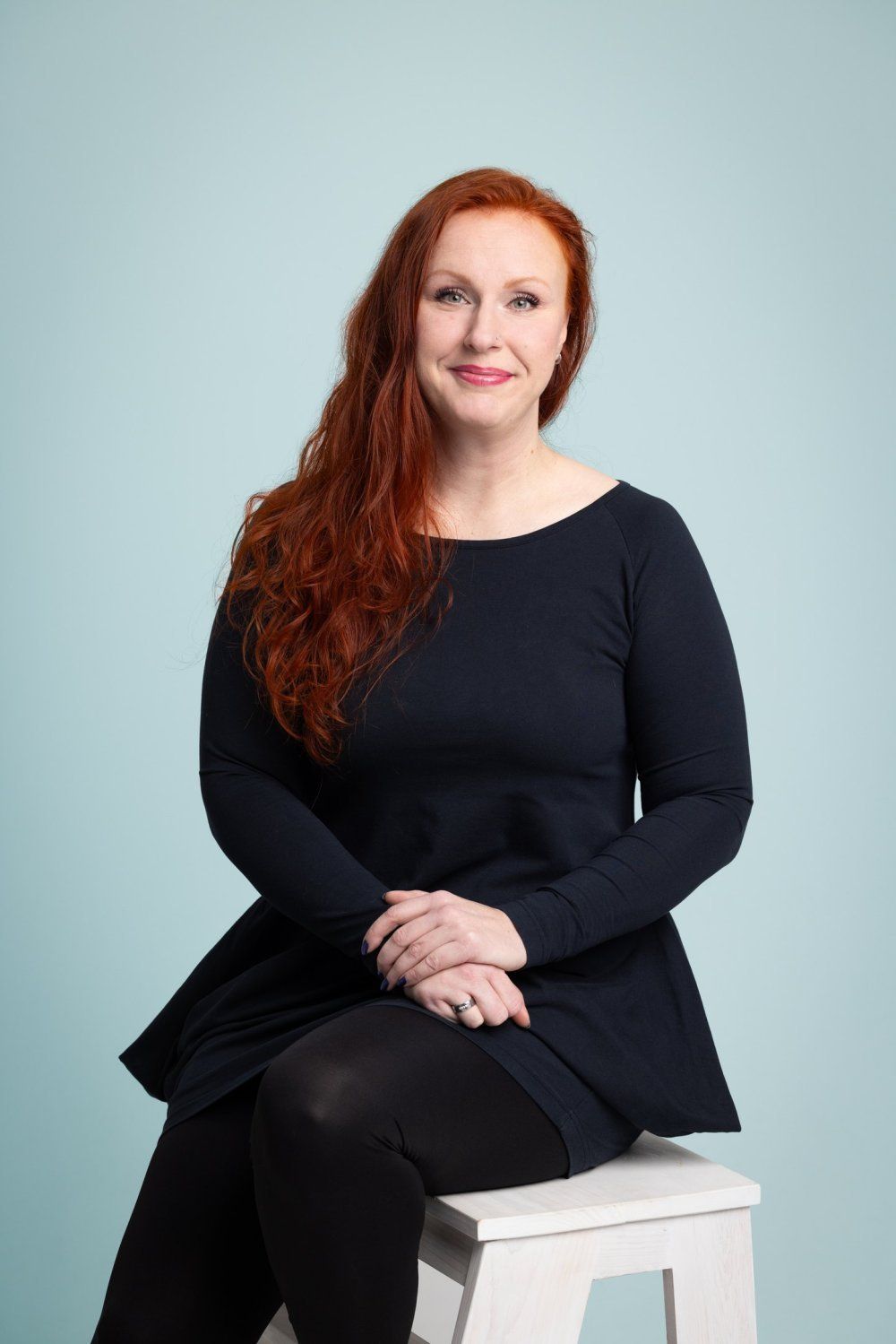 Punatukkainen henkilö mustassa pitkähihaisessa paidassa ja legginsissä istuu valkoisella jakkaralla vaaleansinistä taustaa vasten.