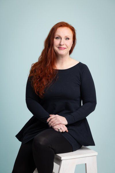 Pitkät punaiset hiukset omaava henkilö, jolla on yllään tumma pitkähihainen paita ja mustat leggingsit, istuu valkoisella jakkaralla sinistä seinää vasten.