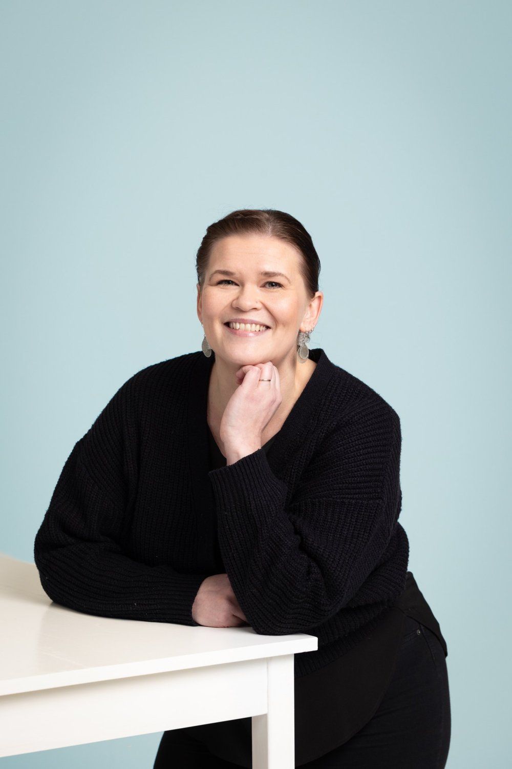 Ruskeatukkainen henkilö mustassa neuleessa hymyilee ja lepää leukaansa kädellään valkoisen pöydän ääressä.