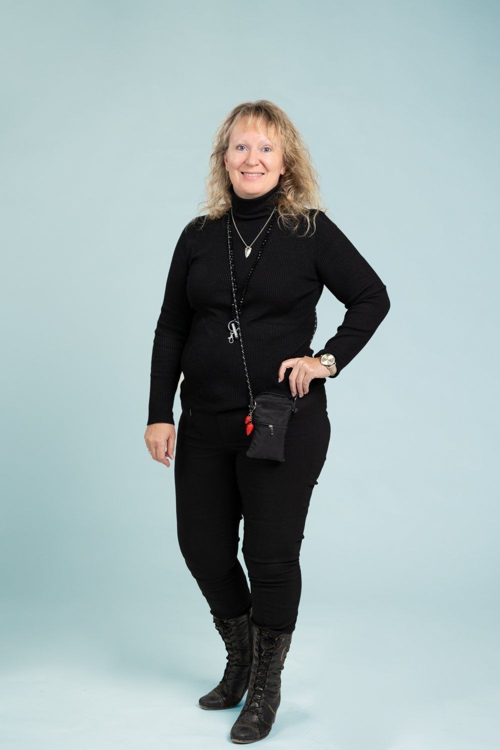 Hymyilevä henkilö seisoo vaaleansinistä taustaa vasten, yllään musta poolopaita, mustat housut ja mustat saappaat.