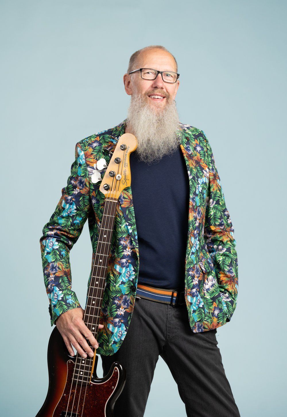 Hymyilevä pitkäpartainen ja silmälasillinen henkilö pitää yllään kukkakuvioista takkia ja pitelee bassokitaraa sinistä taustaa vasten.