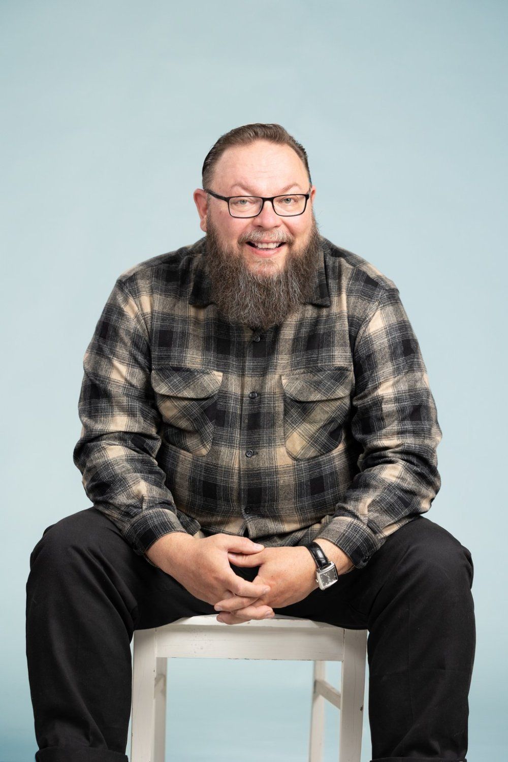 Hymyilevä parrakas ja silmälasipäinen henkilö istuu jakkaralla sinistä taustaa vasten yllään ruutupaita ja mustat housut.